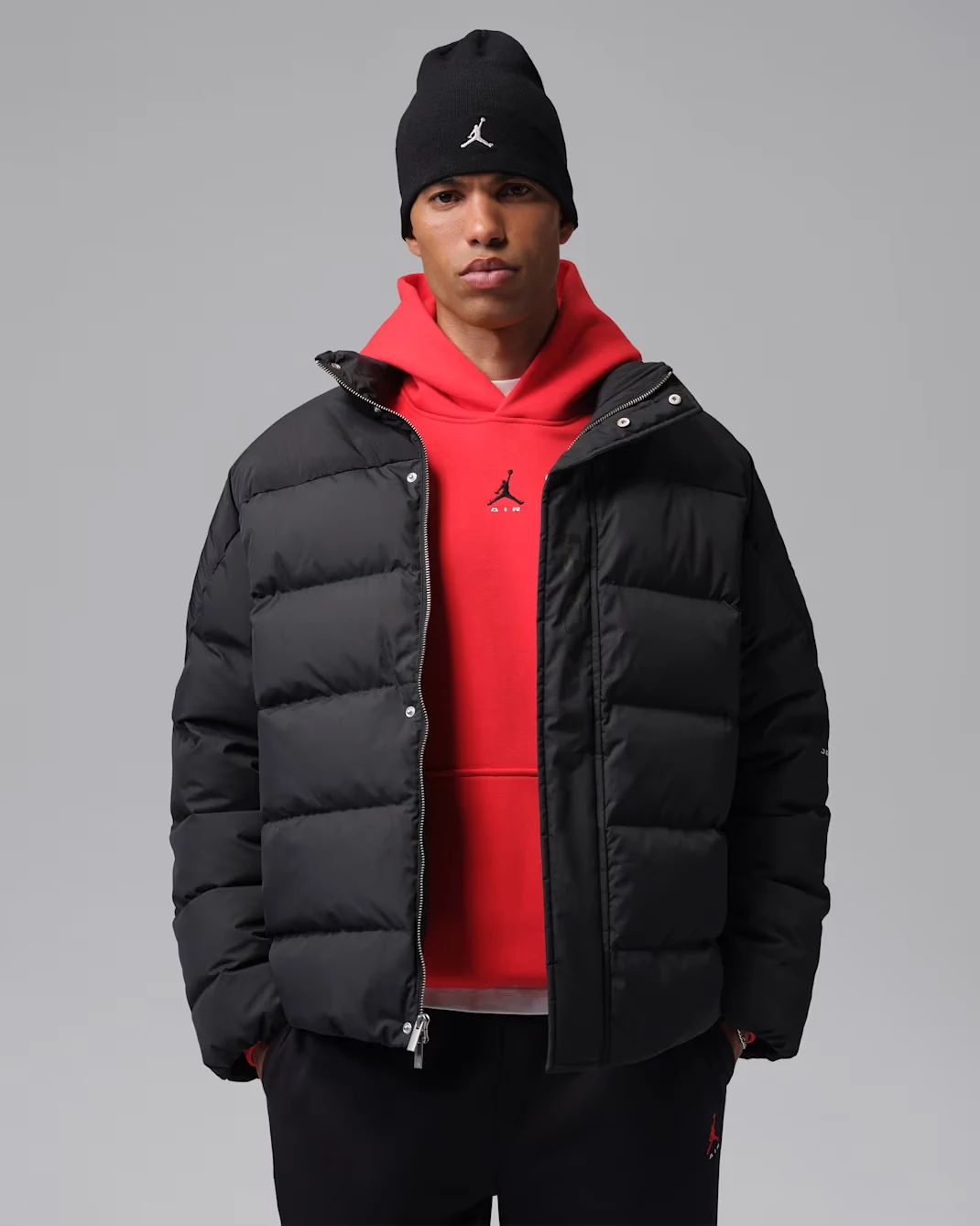 Model wearing a black Jordan Flight down puffer jacket layered over a bright red Jordan hoodie with a black beanie.